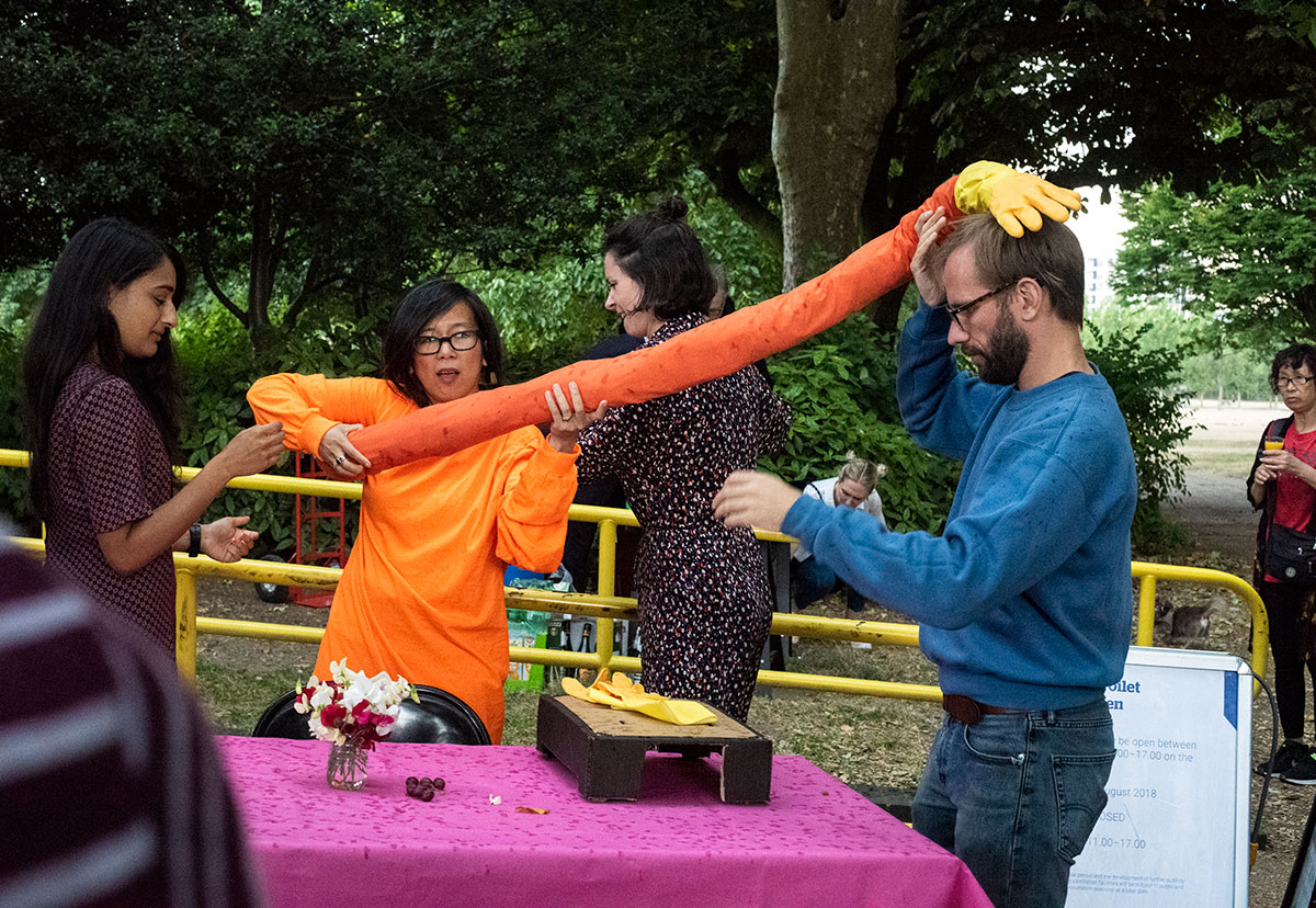 A group of people crowded around a pink table top. The person in the middle is wearing a orange t-shirt and holding a long makeshift arm with a yellow rubber glove on the end which is resting on the head of another person on the right hand side of the image.