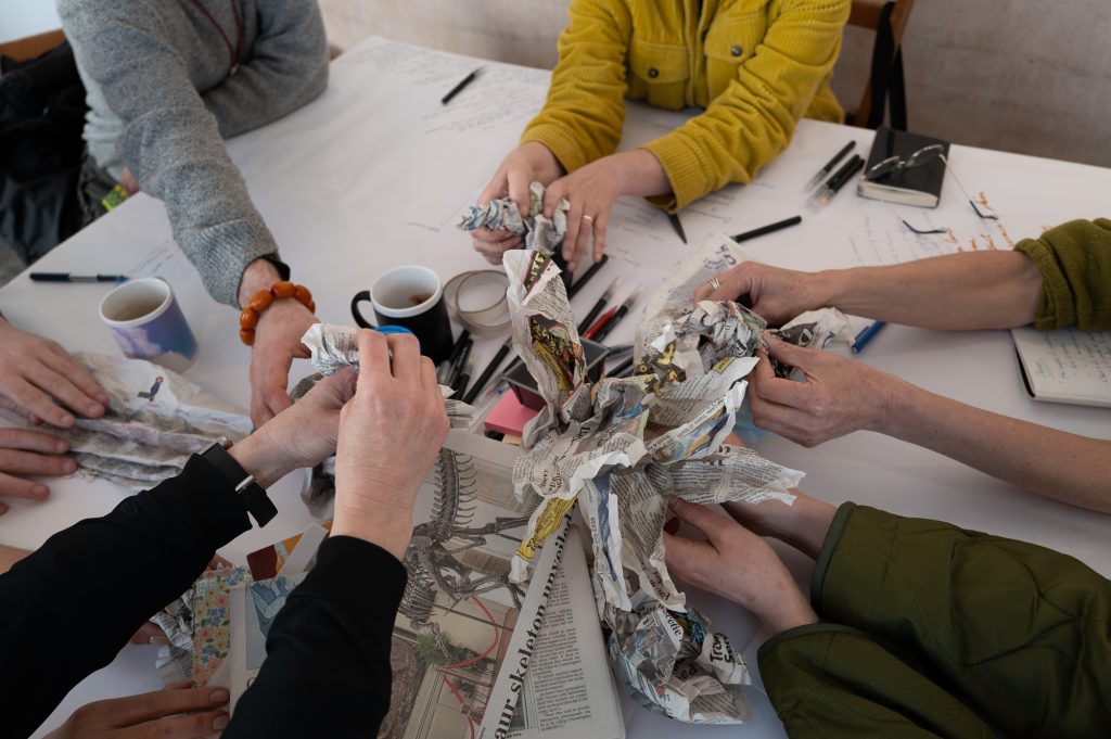 A group of hands around a table holding bits of newspaper fashioned into different shapes, mimicking various different local species we each observed at the seaside before returning to the workshop space.